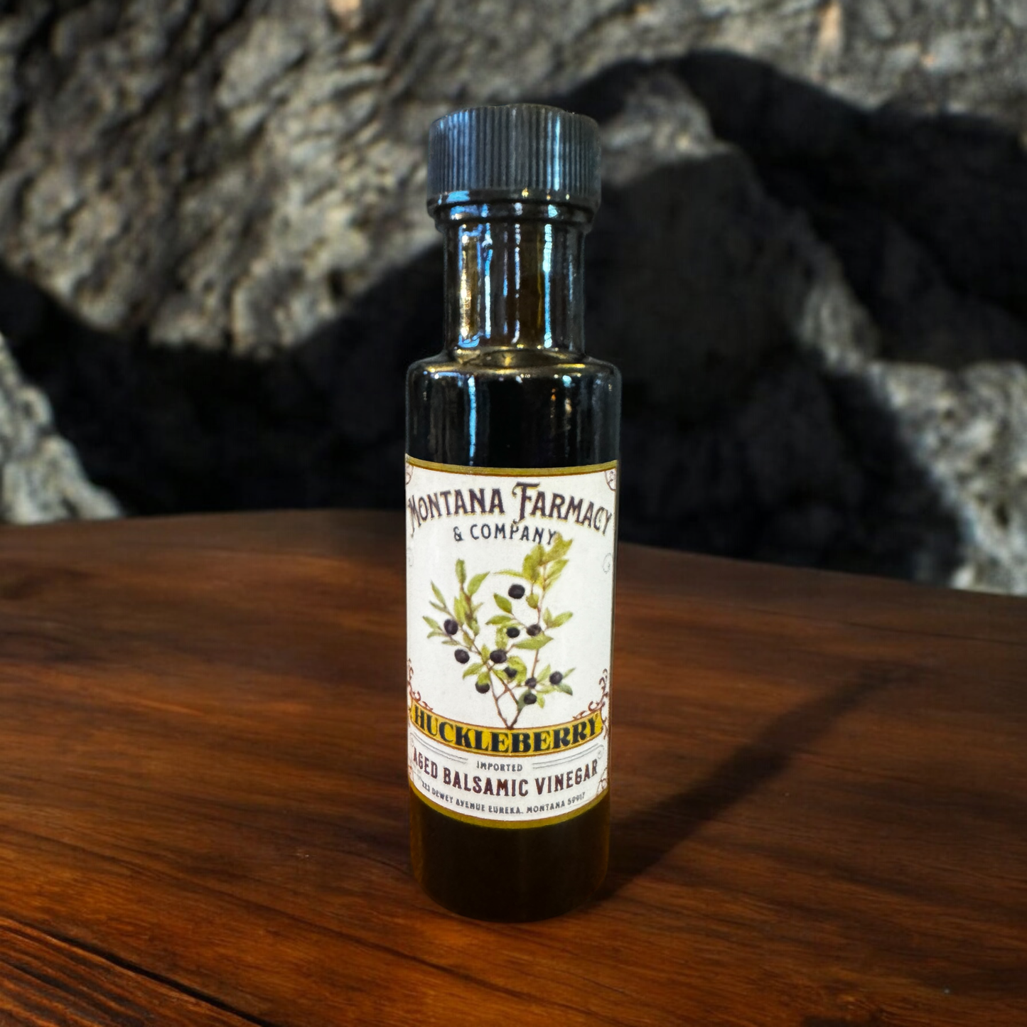 Bottle of Montana Farmacy & Company huckleberry balsamic vinegar on a wooden surface with a textured background.
