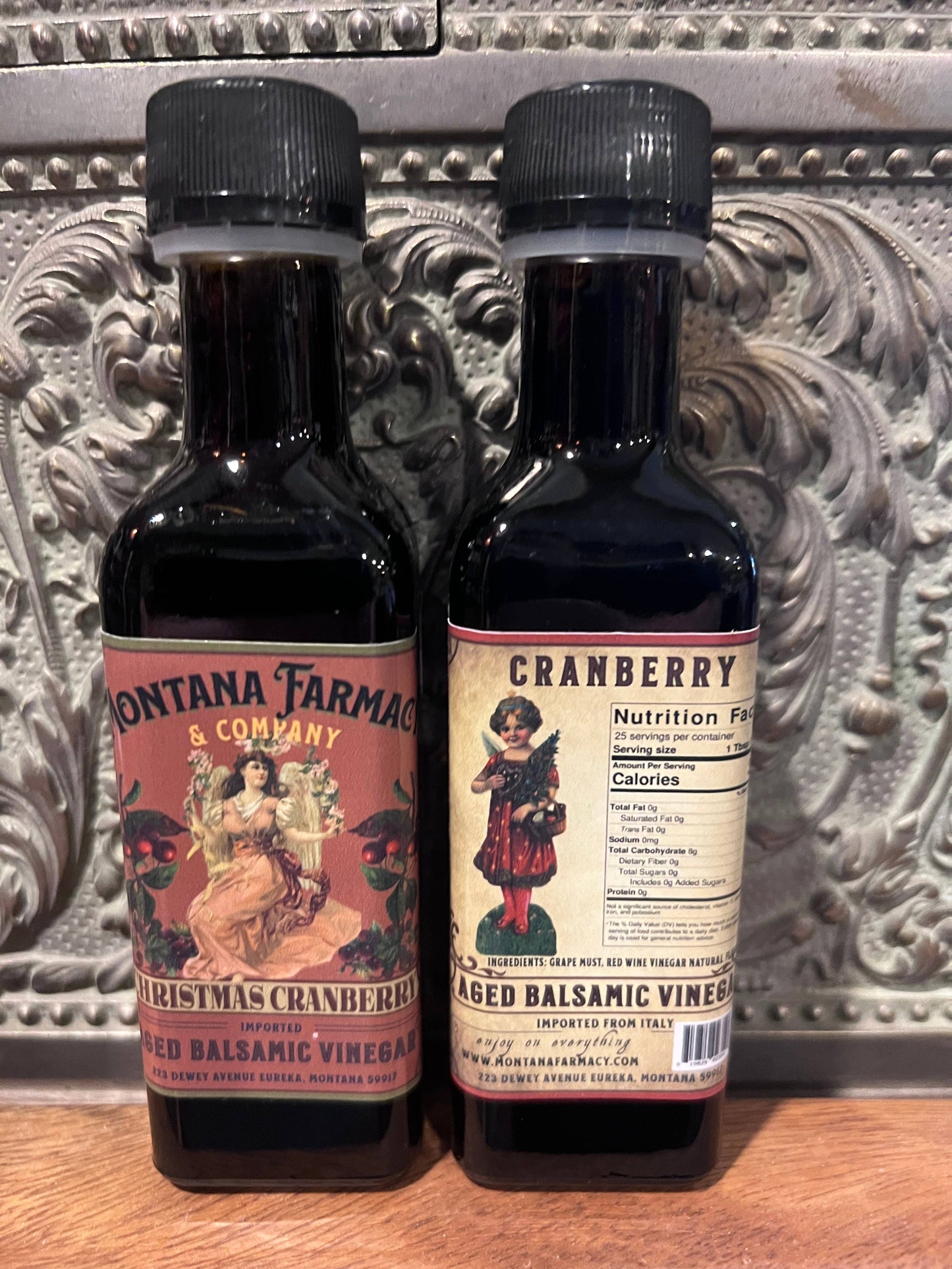 Two bottles (picture of front and one of the back) of Montana Farmacy & Company cranberry aged balsamic vinegar on a decorative surface.