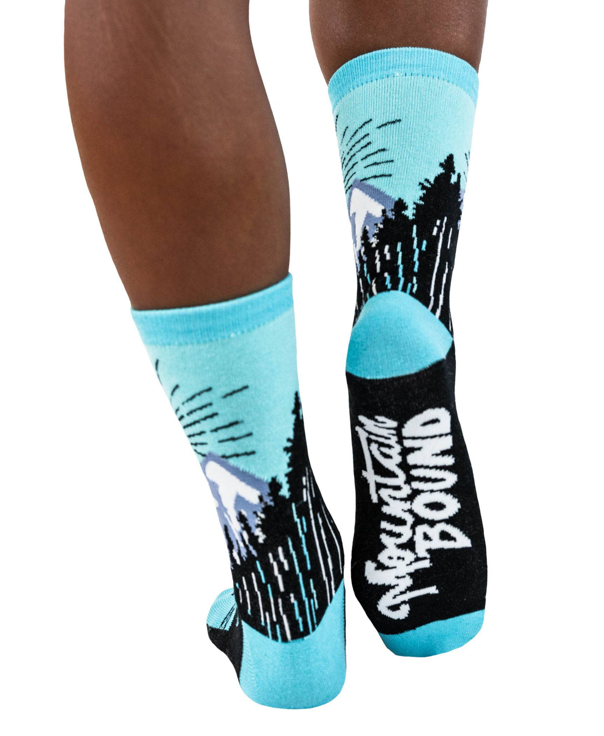 A pair of crew socks worn on a person's legs, featuring a graphic design of pine trees and mountains with the text 'Mountain Bound' in white on a black section, set against a plain white background.