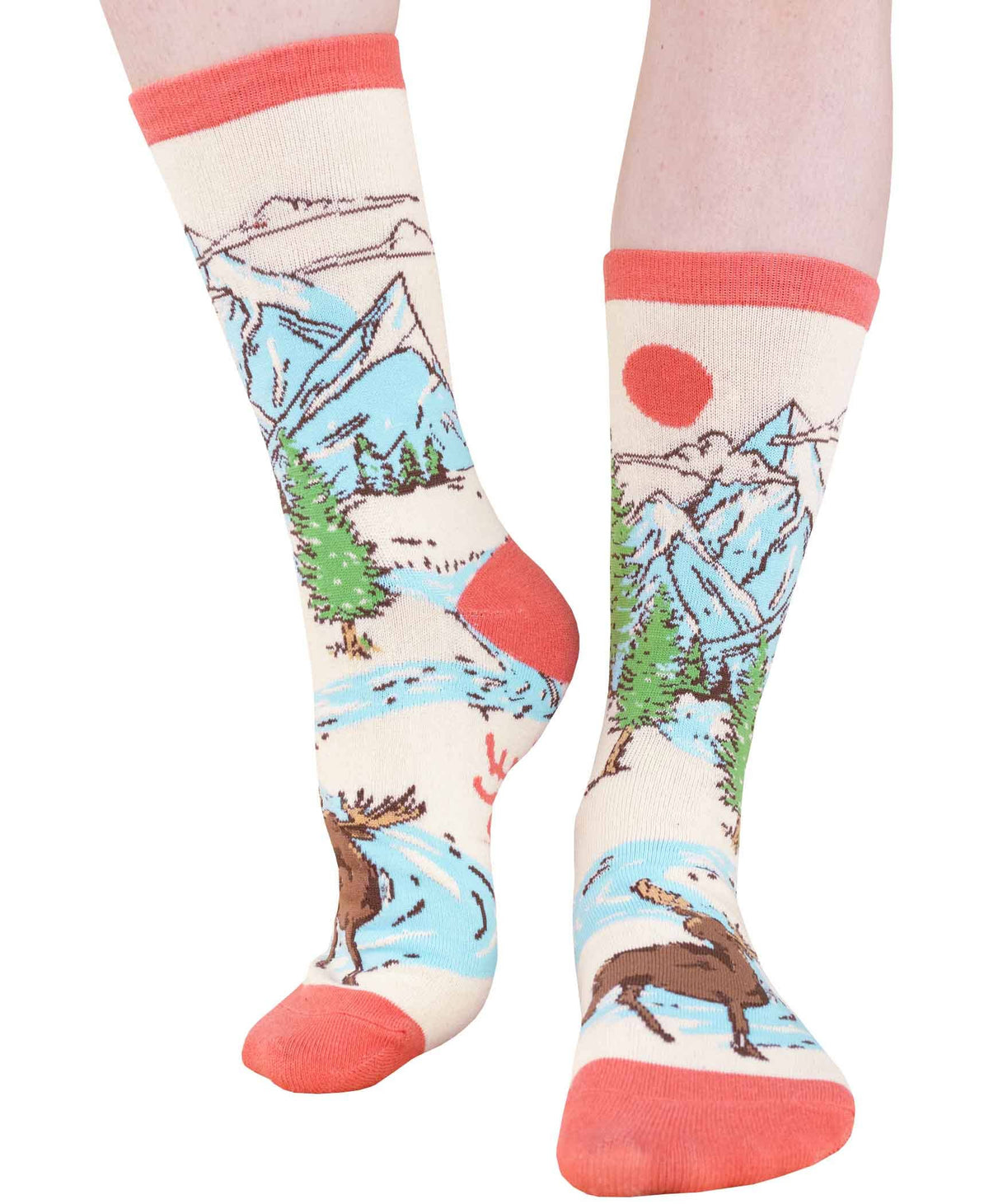 A pair of crew socks worn on lower legs, featuring a scenic landscape print with mountains, trees, a river, a moose, and a red sun on an off-white background with coral pink toe, heel, and cuff details.
