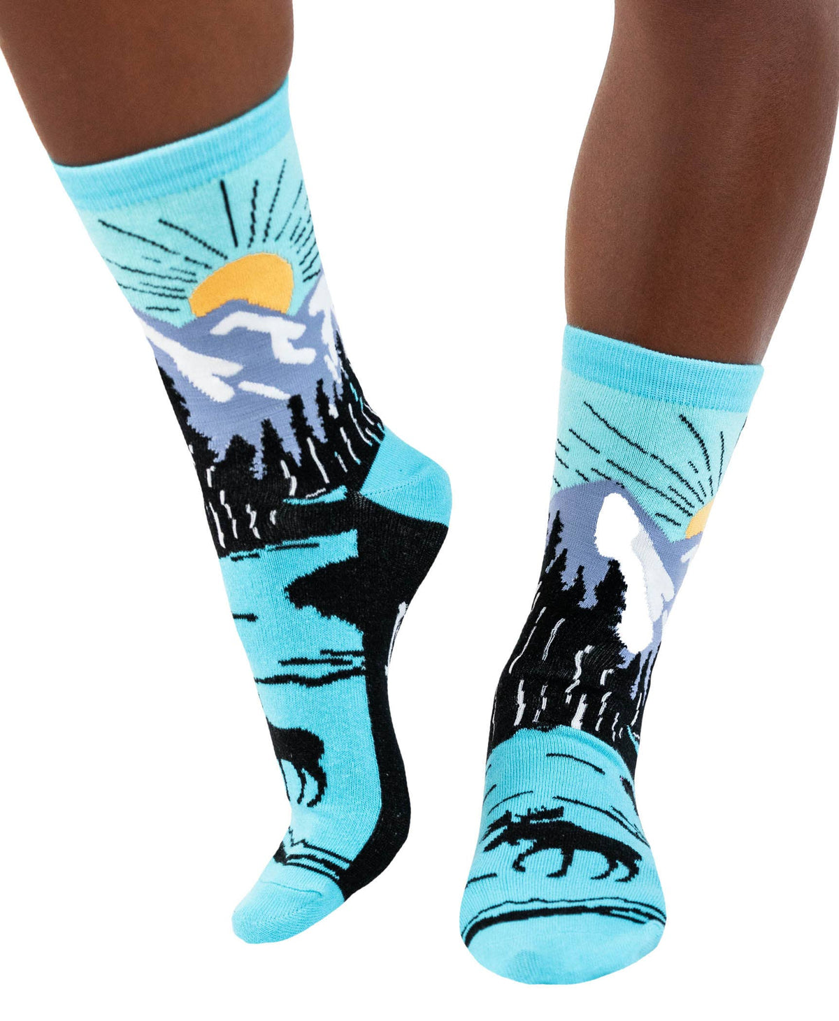 A pair of crew socks worn on legs, featuring a scenic mountain landscape design with a yellow sun, blue sky, black pine trees, and silhouettes of a moose and bear on a white background.